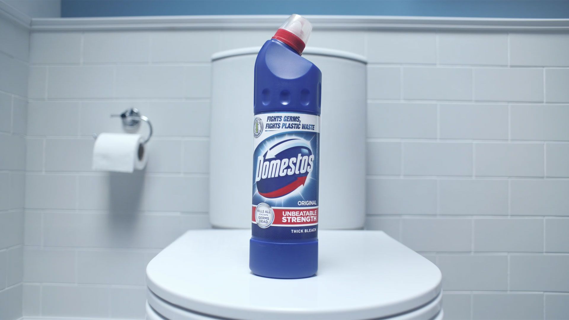 Một chai nước tẩy Domestos Original màu xanh dương được đặt trên nắp bồn cầu màu trắng trong phòng tắm. Chai có vòi phun màu đỏ vát cạnh và nhãn hiệu lớn màu đỏ và xanh dương. Phía sau là những bức tường lát gạch trắng và giá đựng giấy vệ sinh.