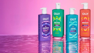 Bốn chai sữa tắm Olly Mood + Skin nhiều màu sắc được đặt trên một bề mặt phản chiếu, phía sau là nền hồng chuyển sắc.